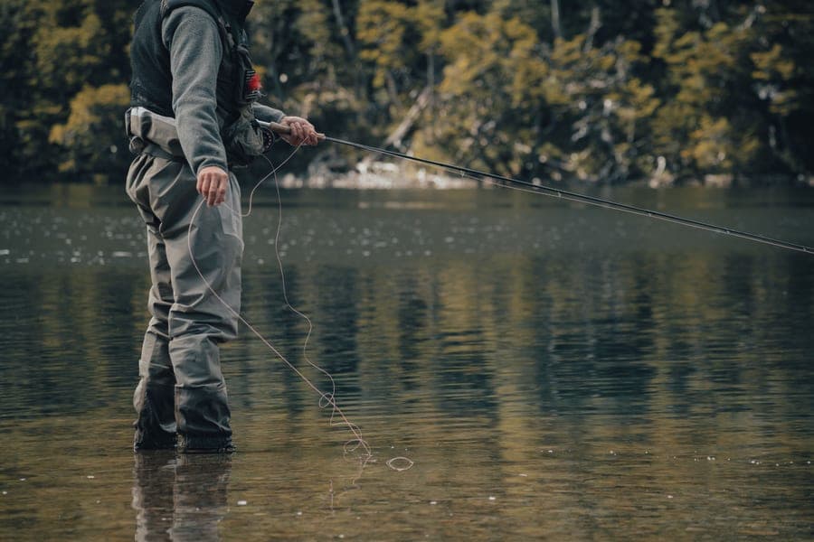 Angler in gray chest waders standing in a calm, tree-lined lake